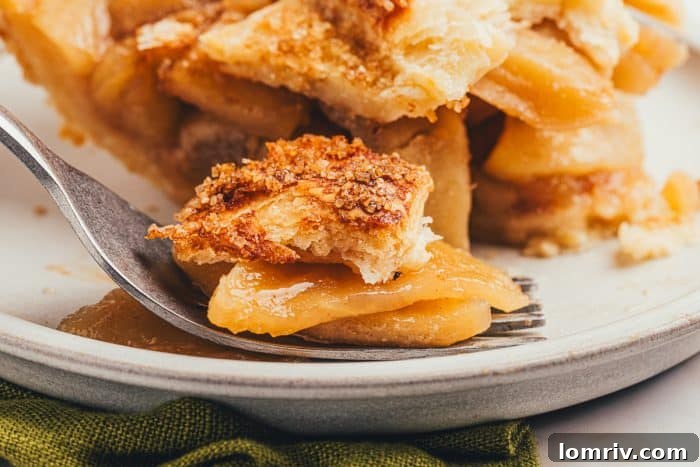 A fork laden with a perfect bite of apple pie rests on a white plate, with a full slice of pie visible in the background.