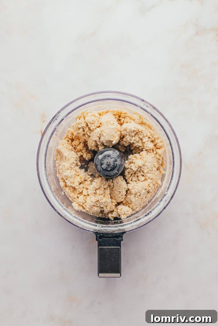 A food processor containing flour, sugar, salt, butter, and grated cheddar cheese, illustrating the mixture before it's pulsed into a coarse meal.