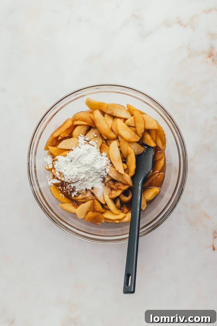 The apple pie filling, now cooled, is in a glass bowl, with cornstarch being sprinkled over it and a black spatula ready to mix.