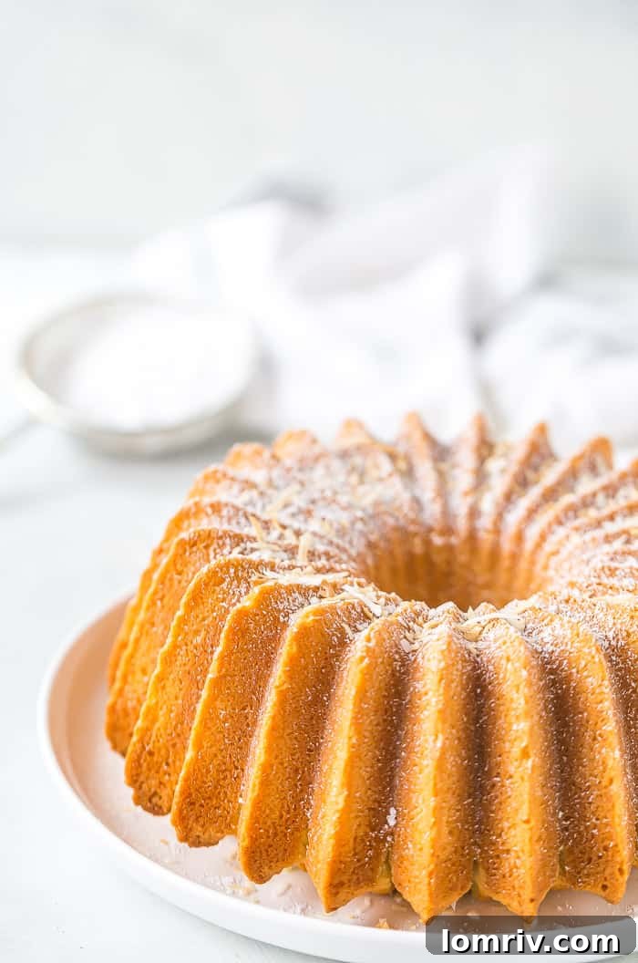 A beautifully baked, golden-brown Coconut Bundt Cake, garnished with a light dusting of powdered sugar, showcased on a white cake stand. Its moist texture and rich coconut flavor are evident.