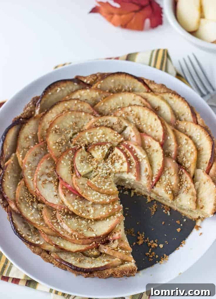 A slice of apple tart with a crumbly sweet crust, a thick layer of frangipane, tender baked apples, and visible sesame seeds, ready for a bite.