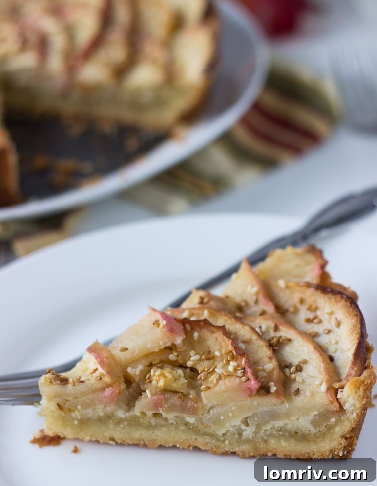 A close-up shot of a golden-brown apple tart with a rich, nutty frangipane layer and visible toasted sesame seeds, served on a light background.