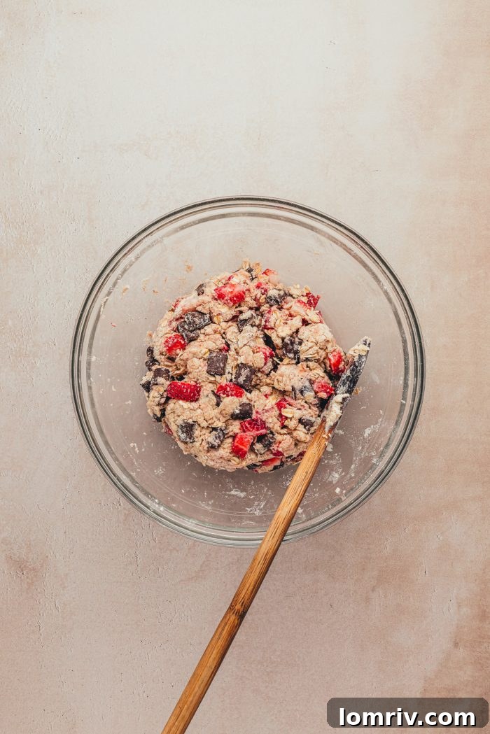 A glass bowl with the dough mixed with strawberries and chocolate and a spatula.