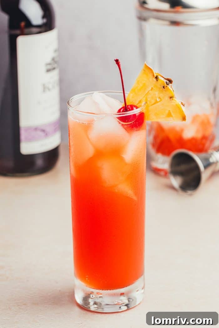 A glass filled with Planter's Punch and decorated with a maraschino cherry and a slice of pineapple with a bottle of rum and a cocktail shaker in the background.