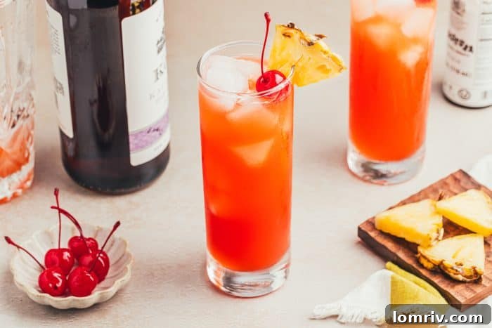 Two glasses filled with Planter's Punch and decorated with maraschino cherries and slices of pineapple with a bottle of rum and a cocktail shaker in the background.