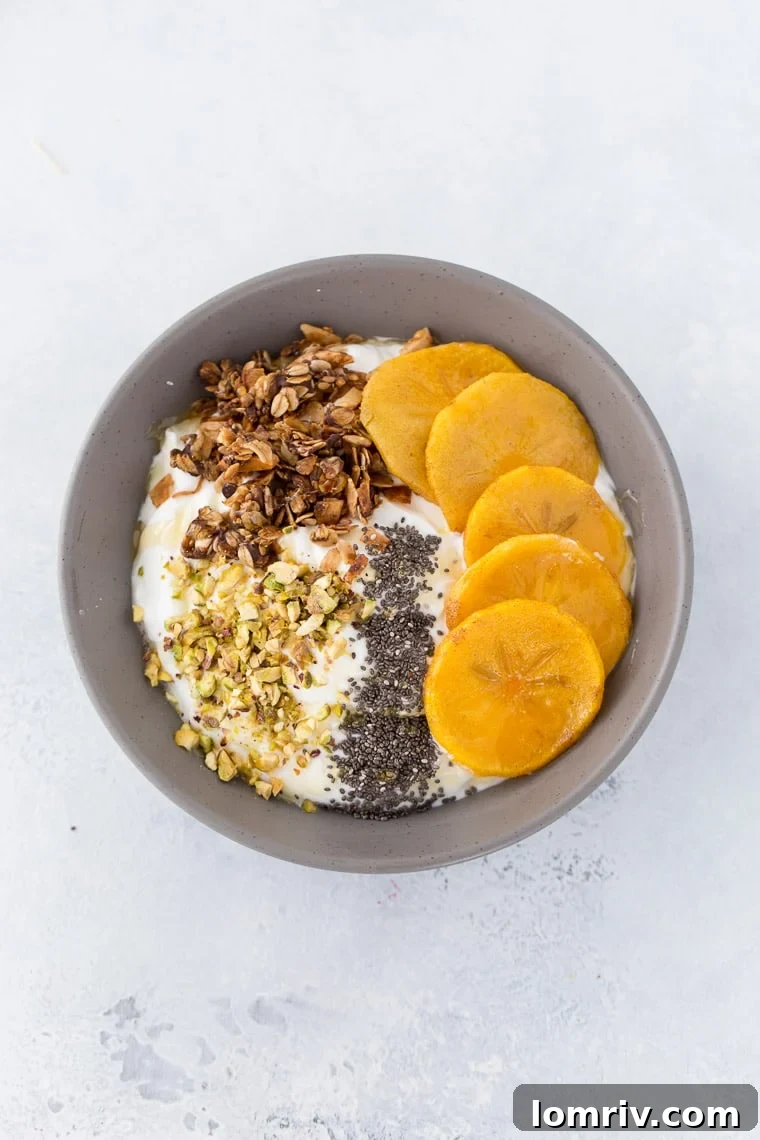 A beautifully arranged roasted persimmon yogurt bowl, featuring vibrant orange persimmons, creamy white yogurt, and crunchy toppings like granola and pistachios.