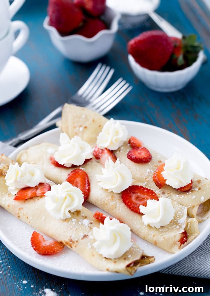 A stack of golden brown crepes ready to be filled, with a bowl of fresh strawberries and mascarpone cream beside them.