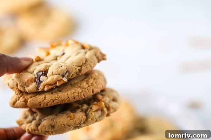 A hand holding three Butterscotch Pretzel Chocolate Chip Cookies, with more cookies blurred in the background, ready to be eaten.