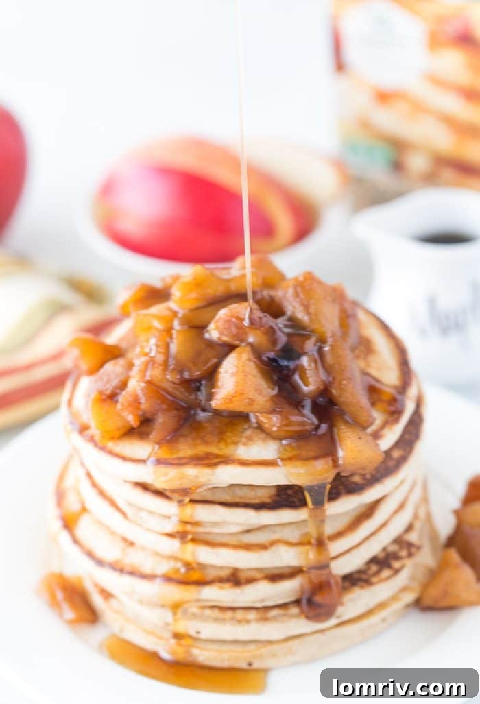 A stack of golden Apple Pie Protein Pancakes with a generous serving of spiced apple topping and a drizzle of maple syrup, presented on a white plate.