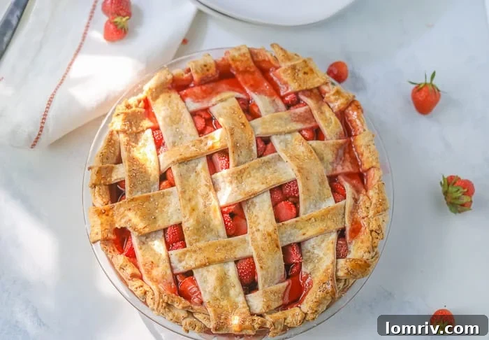 A picture of a homemade strawberry pie with a lattice pie crust and a few fresh strawberries on a table, ready to be served.