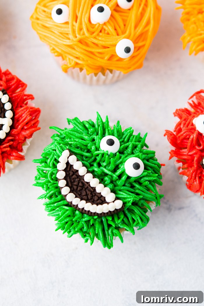 A close-up of a vibrant, fluffy green monster cupcake with two candy eyes and an Oreo cookie mouth showing white frosting teeth, sitting next to a red monster cupcake.