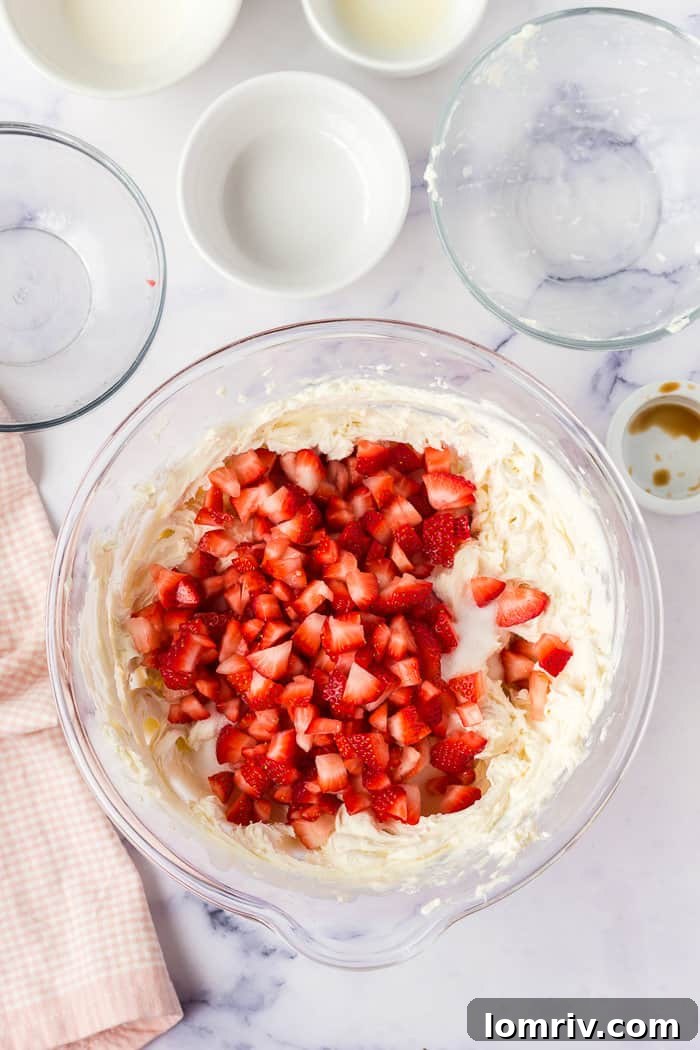 Diced fresh strawberries being gently folded into the creamy cheesecake filling in a large mixing bowl.