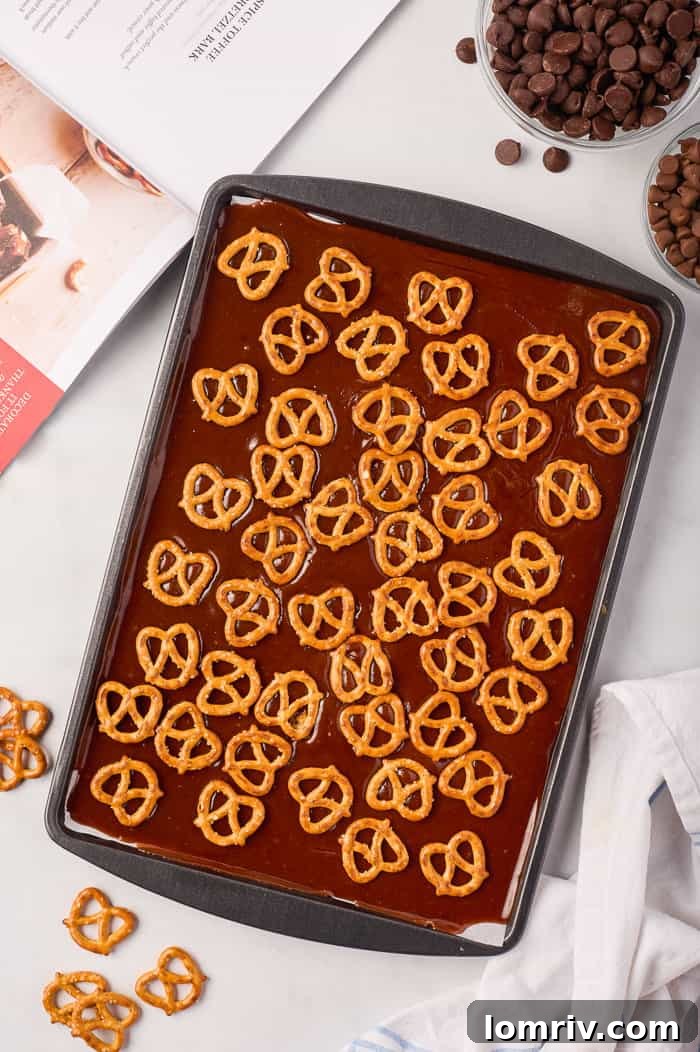 Mini pretzels being pressed into the warm toffee layer on a baking sheet, forming the crunchy base of the pumpkin bark.