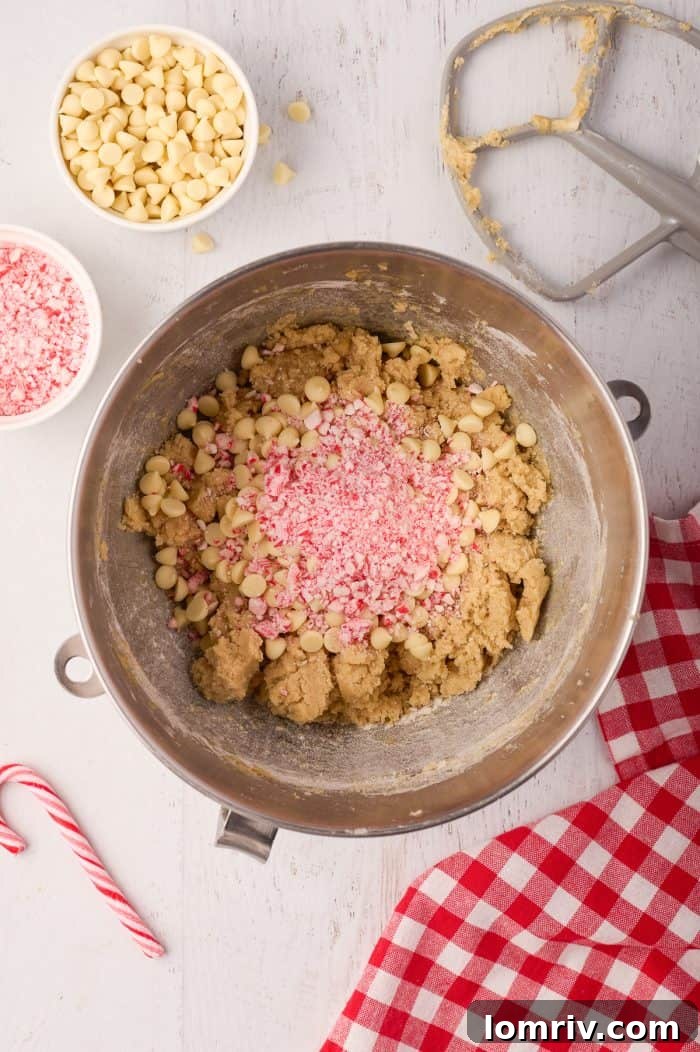 White chocolate chips and crushed candy canes being folded into the prepared cookie dough in a mixing bowl.