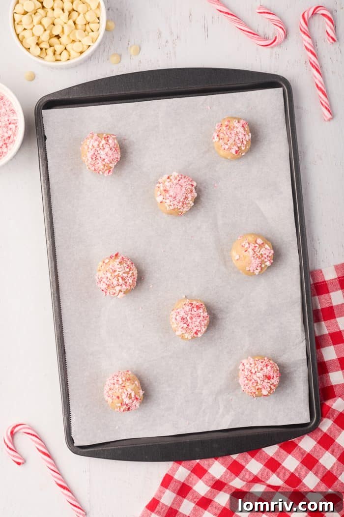 Unbaked peppermint white chocolate cookie dough balls, with some topped with crushed candy canes, neatly arranged on a parchment-lined cookie tray, ready for the oven.