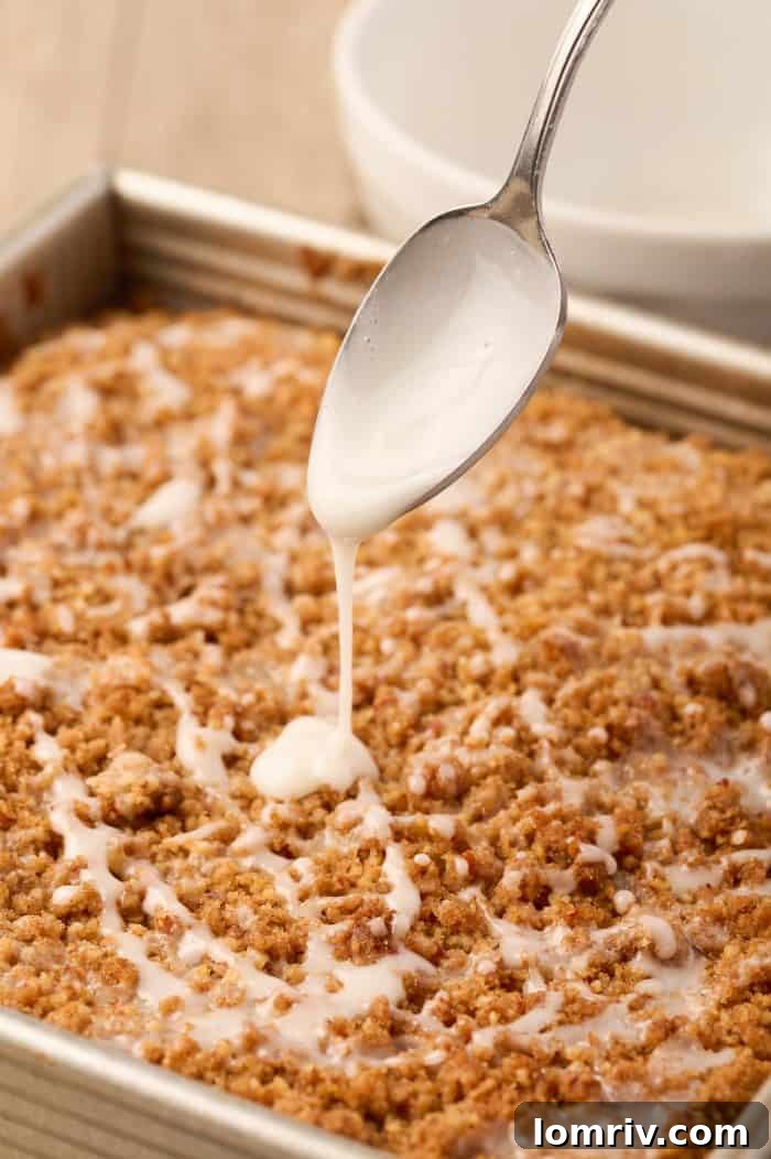 A hand drizzling white icing glaze over the warm sour cream coffee cake in the baking pan.
