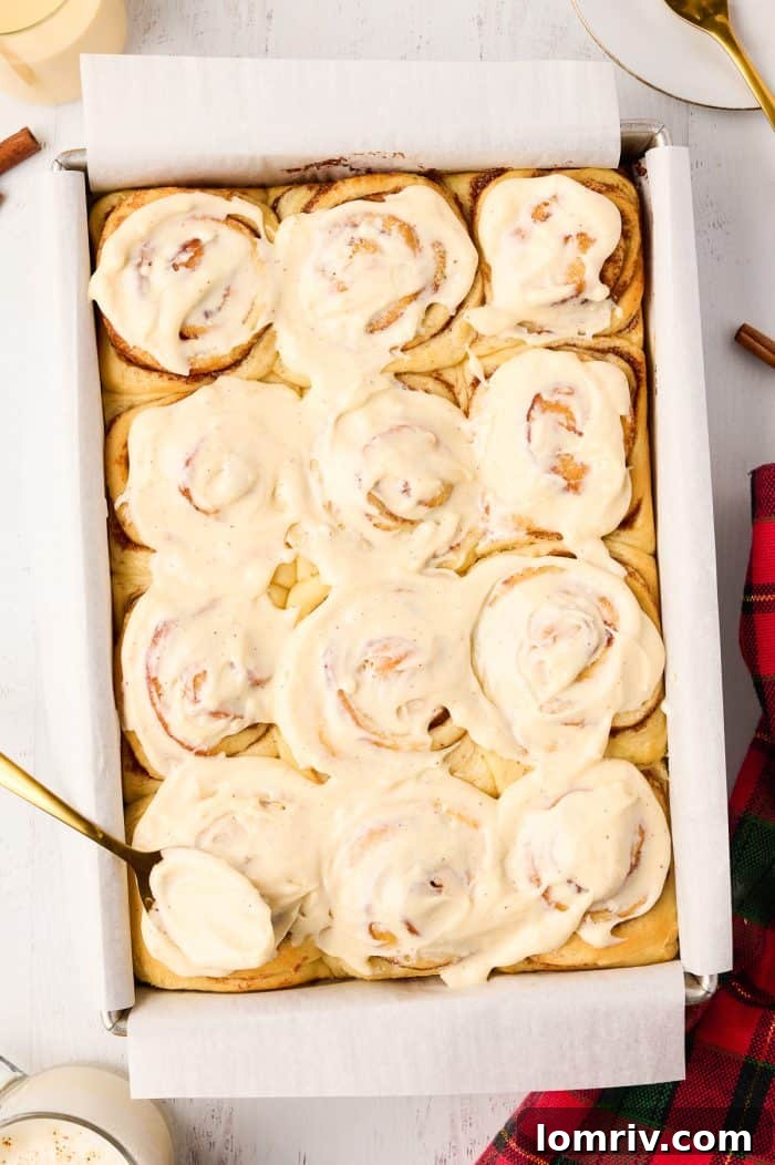 Eggnog cream cheese icing being generously spooned over warm, baked cinnamon rolls.