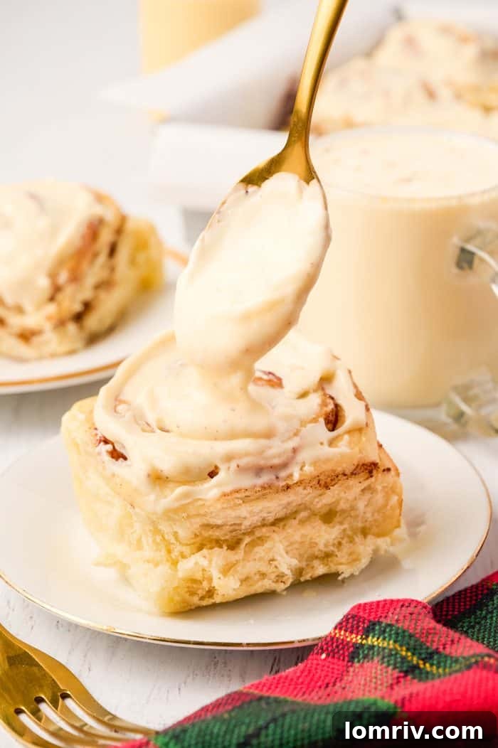 A single Eggnog Cinnamon Roll on a plate with fresh icing being drizzled over it.
