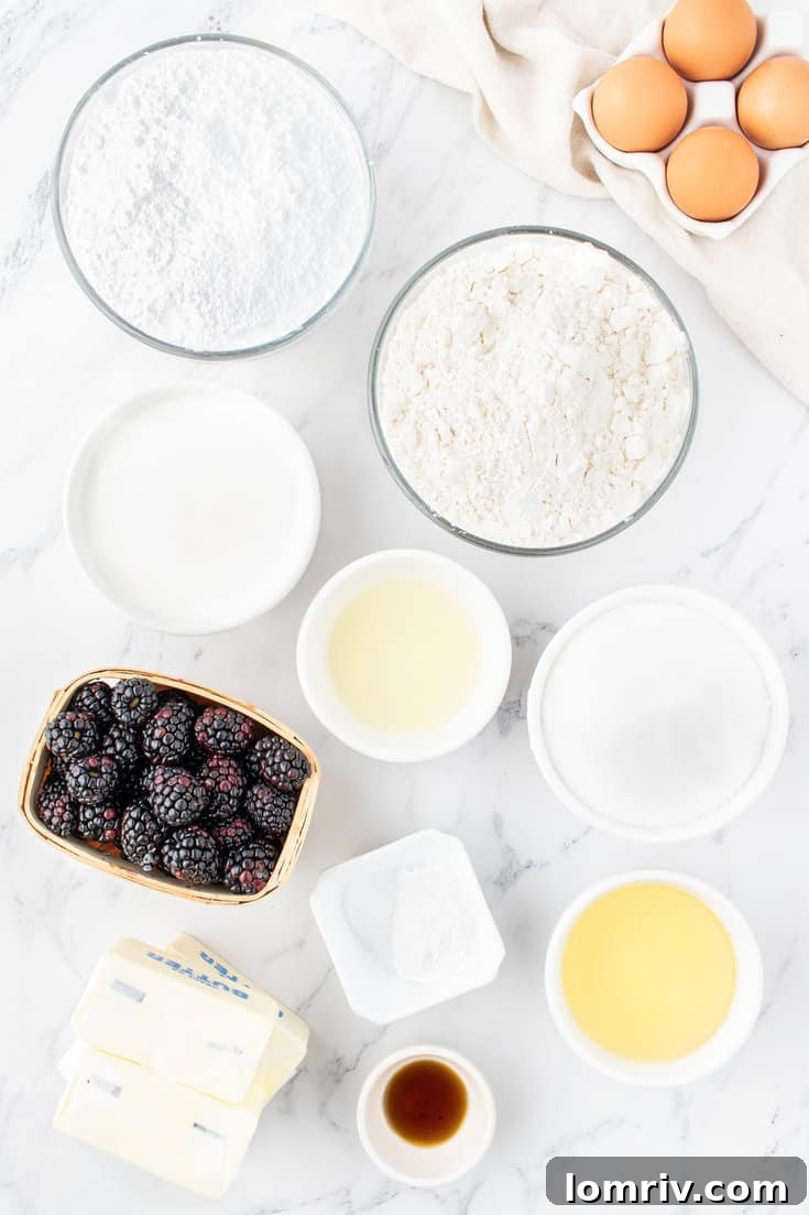 A collection of fresh ingredients laid out on a table, including flour, butter, eggs, milk, sugar, and fresh blackberries.