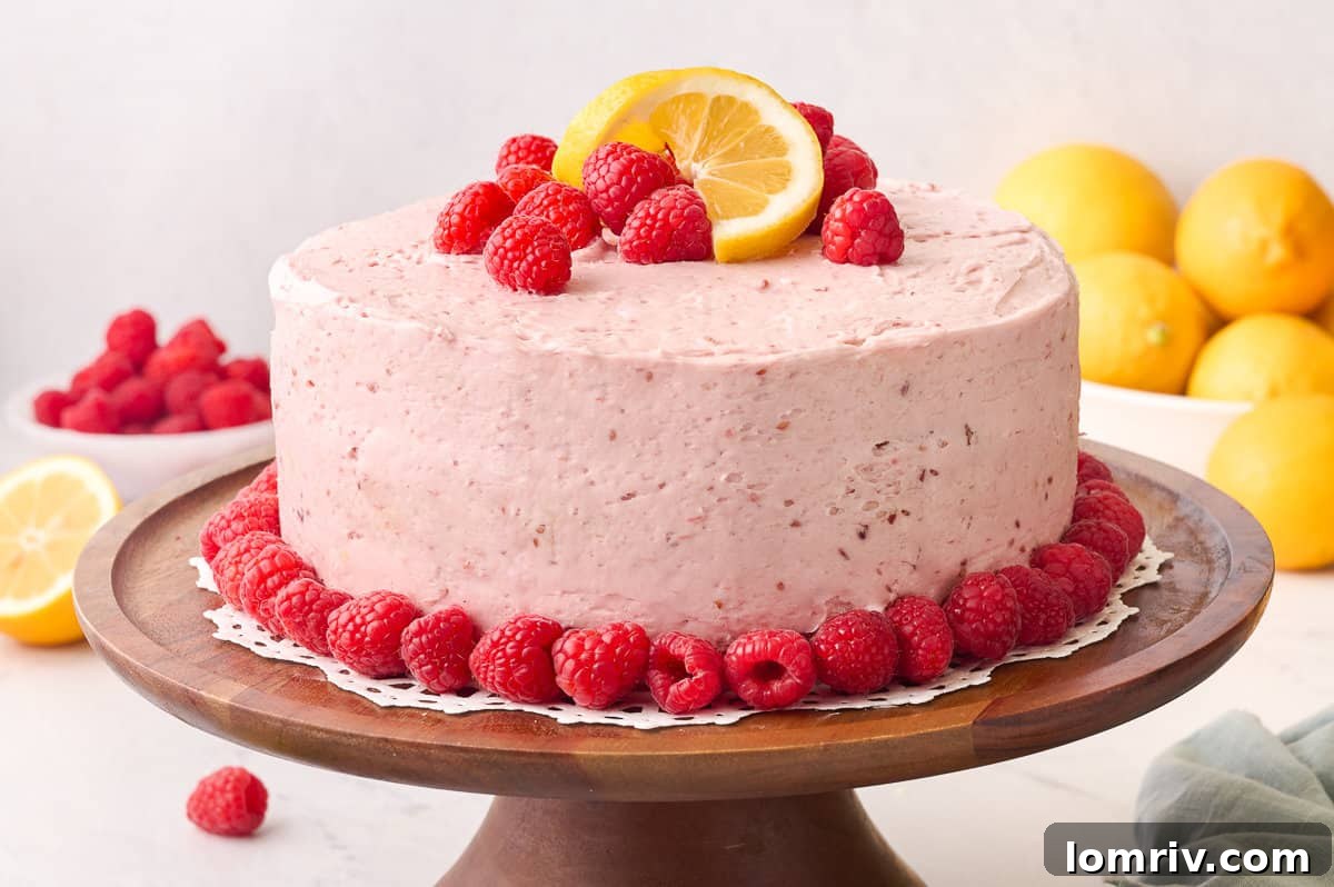 A stunning Lemon Raspberry Layer Cake, beautifully frosted with raspberry buttercream and garnished with fresh raspberries and lemon slices, presented on a white cake stand.