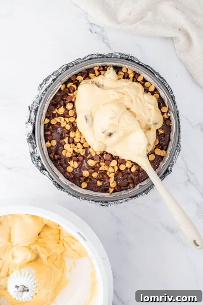 The rich peanut butter cheesecake filling being spooned generously over the cooled brownie crust, creating the next delicious layer of the cheesecake.