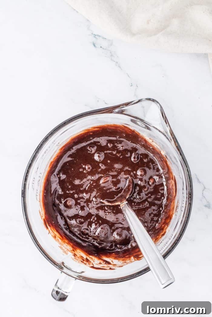 The prepared brownie batter being poured into a 9-inch springform pan lined with parchment paper, ready for baking to form the cheesecake crust.