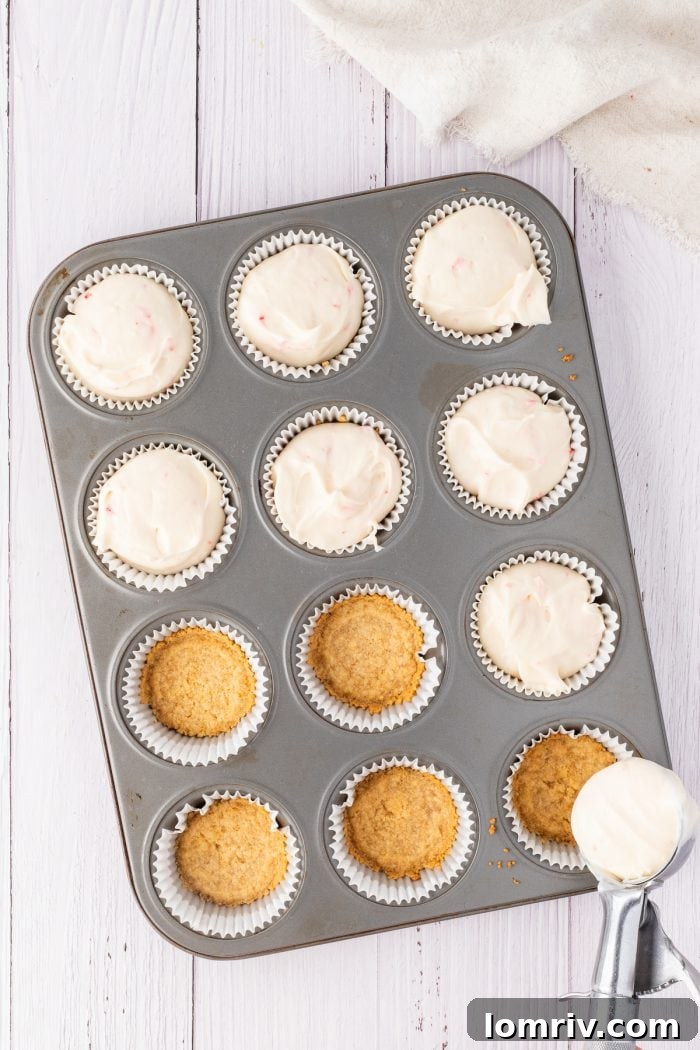 A generous spoonful of creamy cheesecake filling is carefully added on top of a prepared cookie crust in a cupcake liner, building the layers of the dessert.