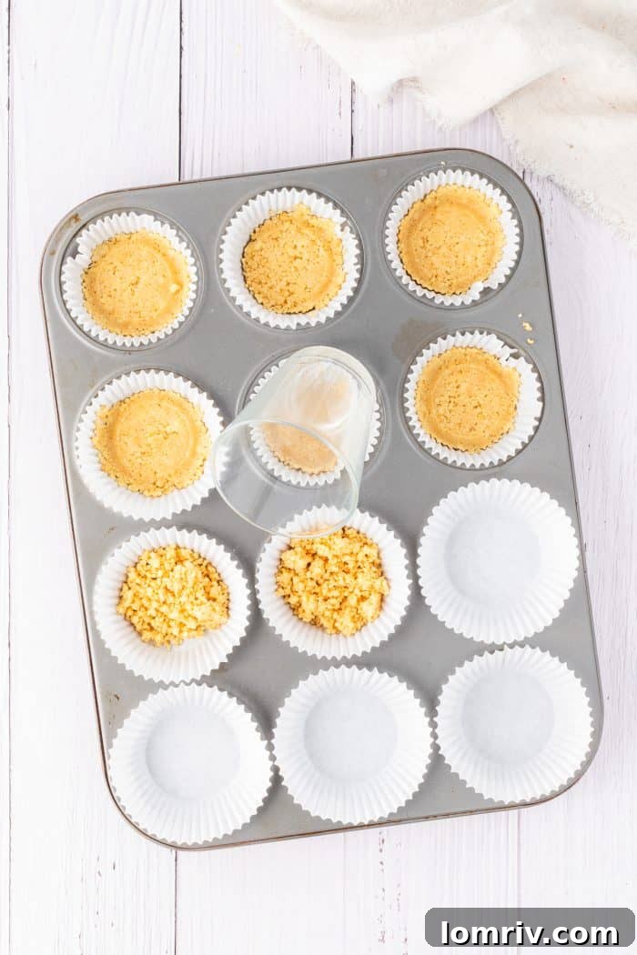 A hand firmly presses down the golden Oreo crust mixture into a cupcake liner using the back of a spoon, ensuring a compact and even base.