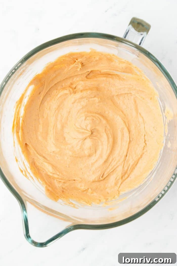 The cream cheese mixture, now blended with vibrant pumpkin puree and aromatic pumpkin pie spice, swirling in a glass mixing bowl.