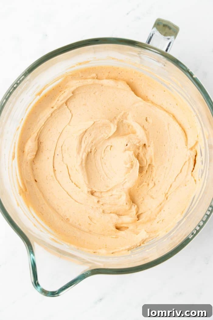 A stream of cold heavy cream being poured into the rich pumpkin cream cheese mixture within a glass mixing bowl, anticipating its transformation into a thick filling.