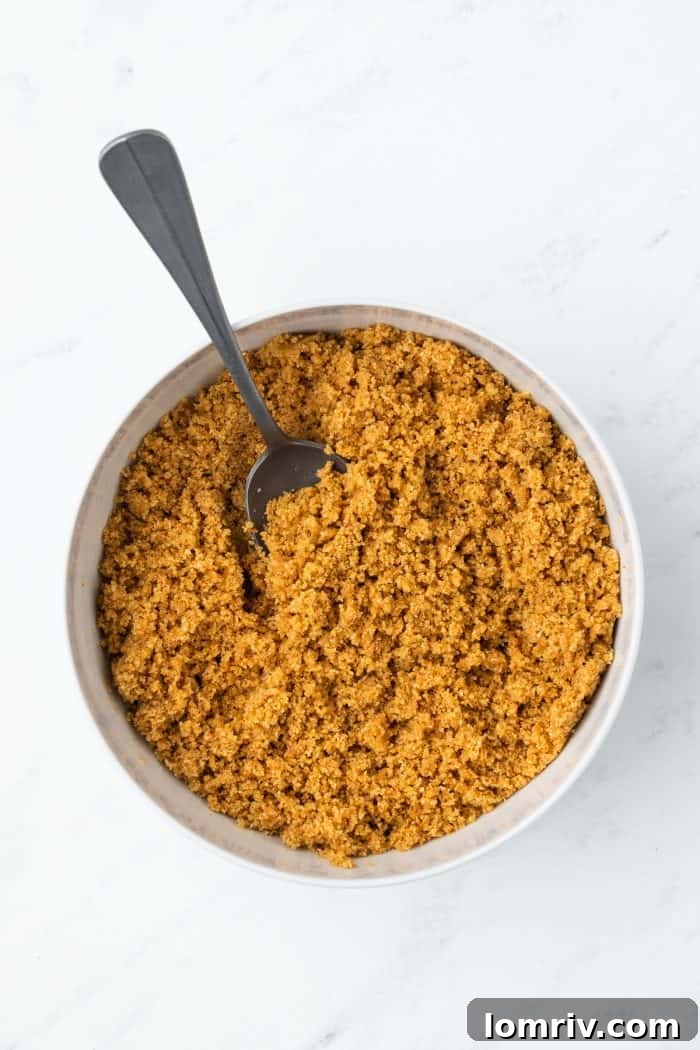 A close-up of graham cracker crumbs thoroughly mixed with melted butter in a transparent glass bowl, ready for the next step.