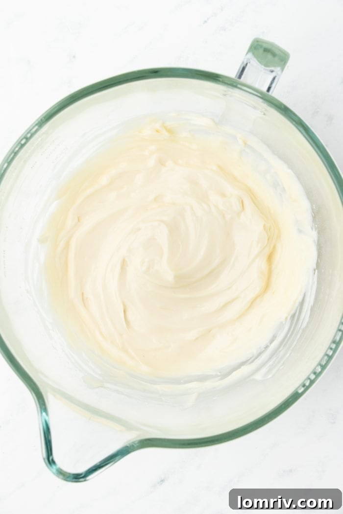 Softened cream cheese sitting in a large glass mixing bowl, poised for the whisk attachment of an electric mixer.