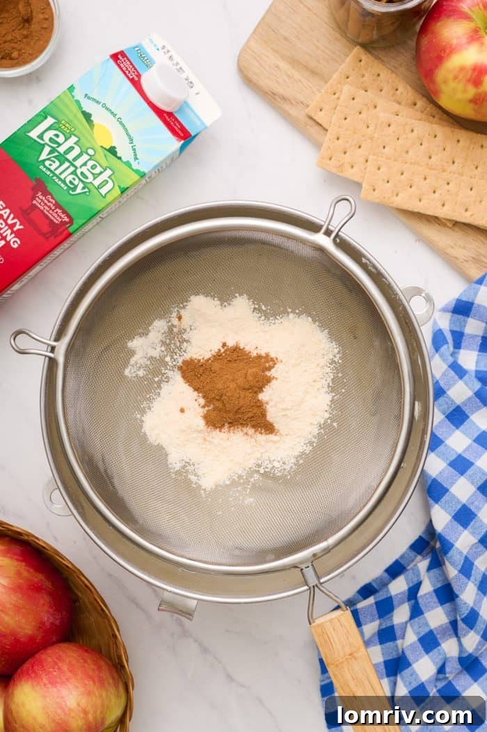 A sieve holding crushed freeze-dried apples and apple pie spice, poised above a bowl of cream cheese mixture.