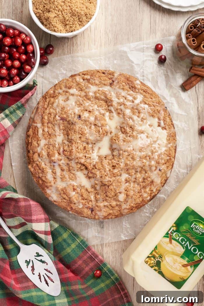 An overview picture of the cranberry eggnog coffee cake with a bowl of cranberries, a cake server, and a bottle of eggnog, set against a festive holiday backdrop.