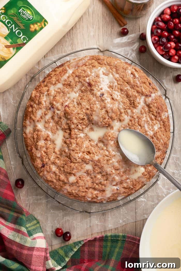 The beautiful moment of drizzling the delicate eggnog icing over the cooled cranberry eggnog coffee cake, creating elegant streaks on the golden surface.