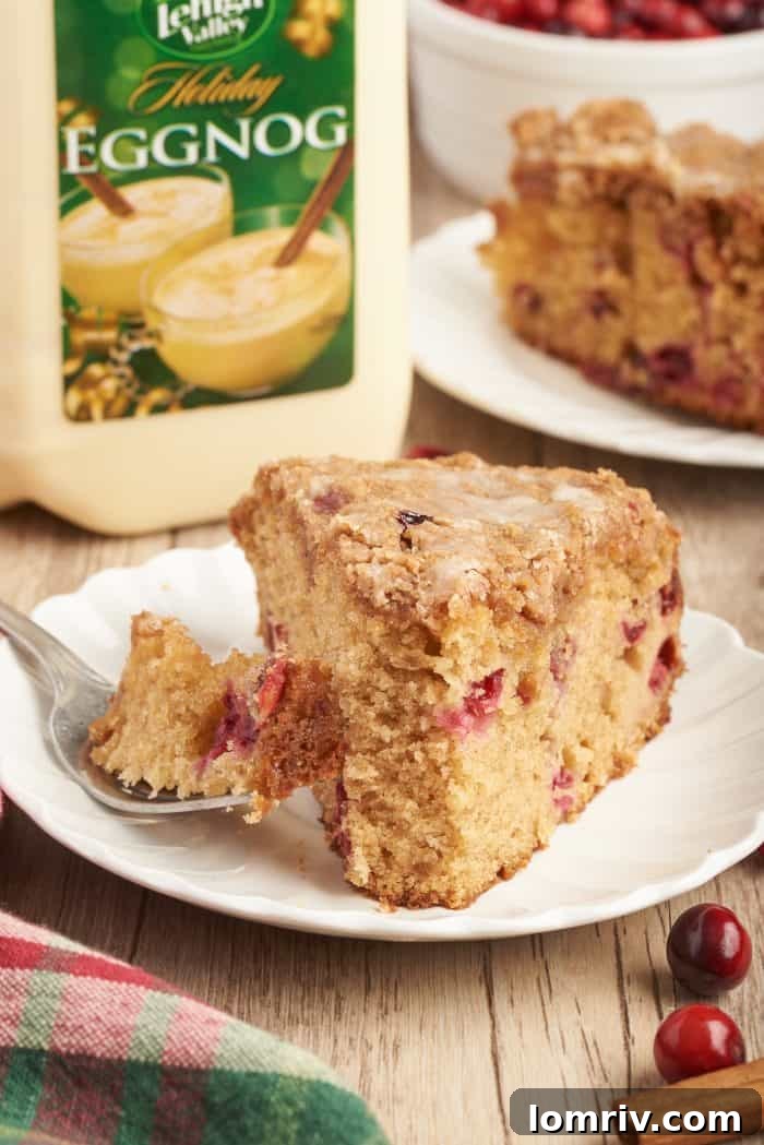 A close-up shot of a slice of cranberry eggnog coffee cake on a white plate, with a fork holding a small bite, highlighting its texture and inviting appearance.