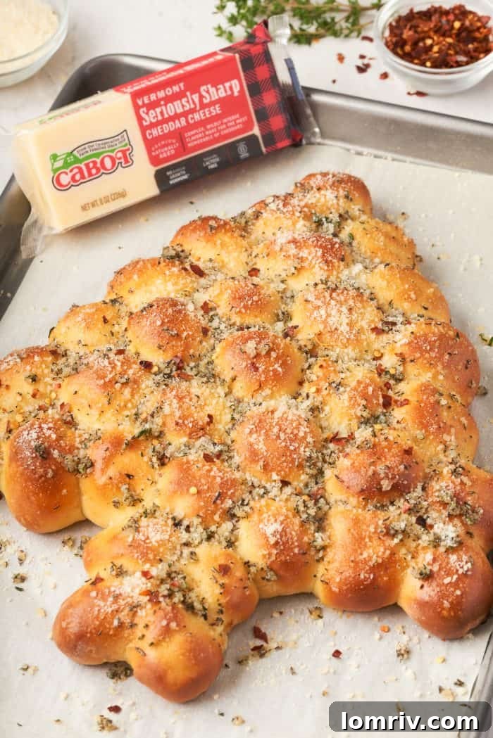 A delectable Christmas tree pull-apart bread, golden brown and garnished with herbs, on a baking sheet, with a block of sharp cheddar cheese beside it.
