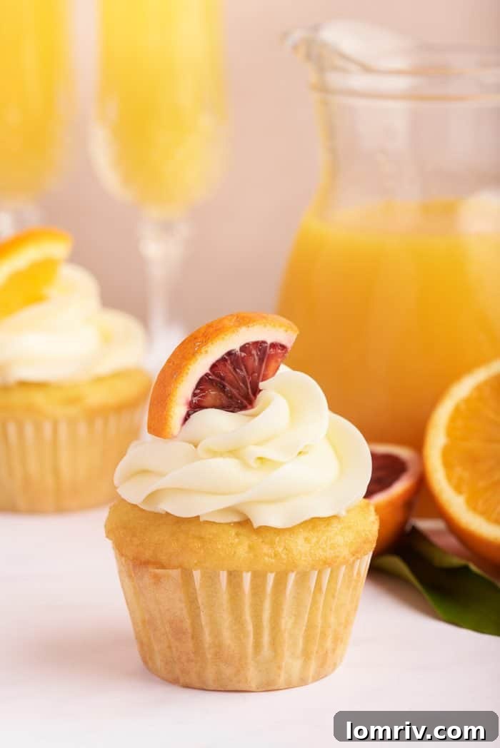 A single mimosa cupcake, beautifully frosted with orange-infused Swiss meringue buttercream and garnished with an orange segment, rests beside a tall glass filled with a bubbly mimosa. In the background, a bottle of fresh orange juice hints at the drink's key ingredient.