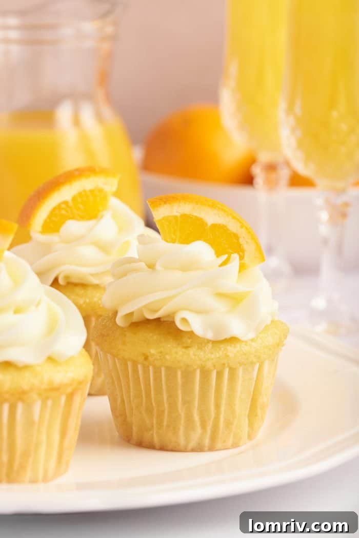 Several mimosa cupcakes are arranged on a pristine white plate, each adorned with a swirl of orange Swiss meringue buttercream and an orange slice. The setting includes a glass of mimosa and a bottle of orange juice, enhancing the thematic presentation.