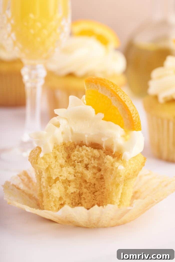 A mimosa cupcake, with a delightful bite taken out of it, reveals its moist, light texture and citrus-infused interior. It is artfully placed next to a glass of mimosa, suggesting a harmonious pairing.