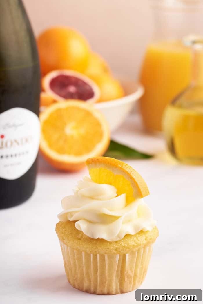 A close-up view of a perfectly frosted mimosa cupcake, adorned with a delicate slice of orange. In the background, a shimmering bottle of Prosecco stands, emphasizing the champagne inspiration behind the dessert.