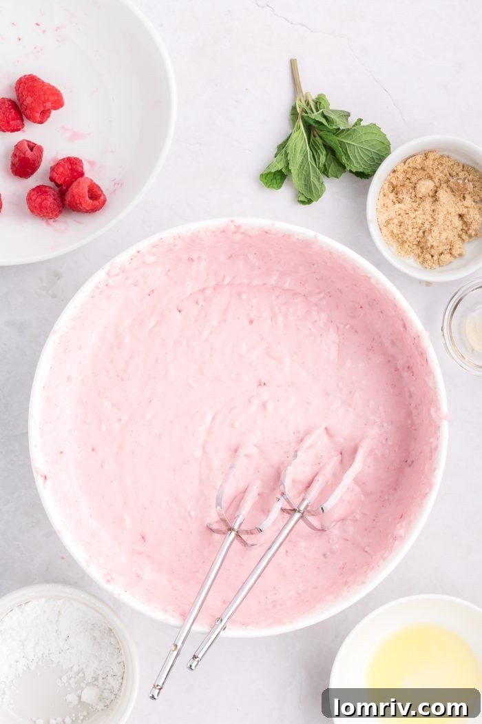 Cream cheese, raspberries, and heavy whipping cream being mixed in a white bowl with a hand mixer.