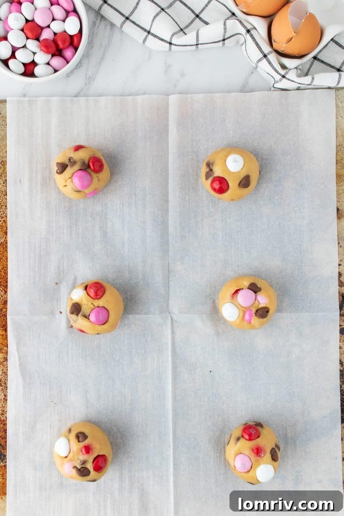 A baking sheet prepared with parchment paper, holding evenly spaced cookie dough balls, each topped with extra M&M's, poised for baking.