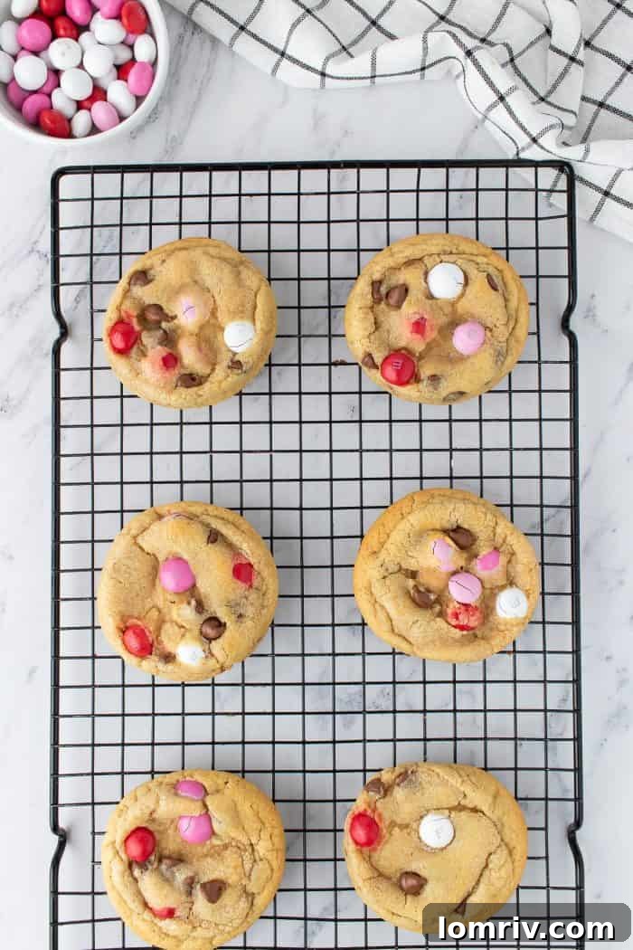 Six freshly baked Valentine's M&M cookies arranged on a wire cooling rack, with a bowl of colorful M&M candies visible in the soft background.