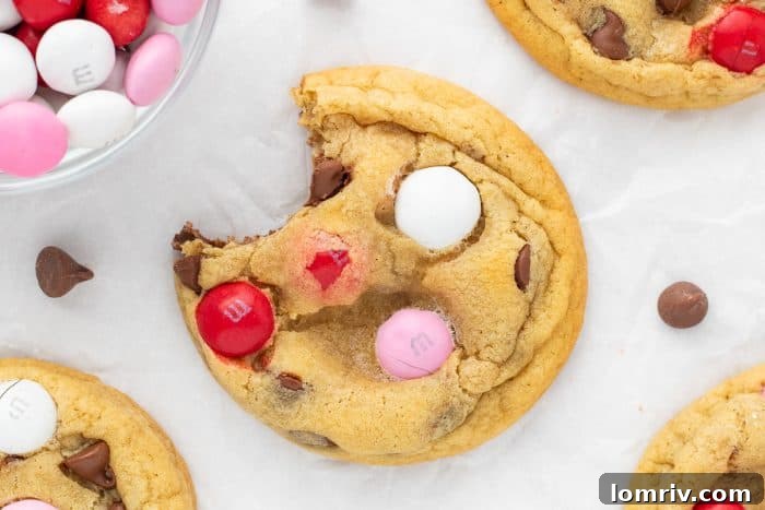 A captivating close-up of a Valentine's M&M cookie, featuring a perfect bite revealing its soft texture and abundant chocolate, with additional cookies artfully blurred in the background.
