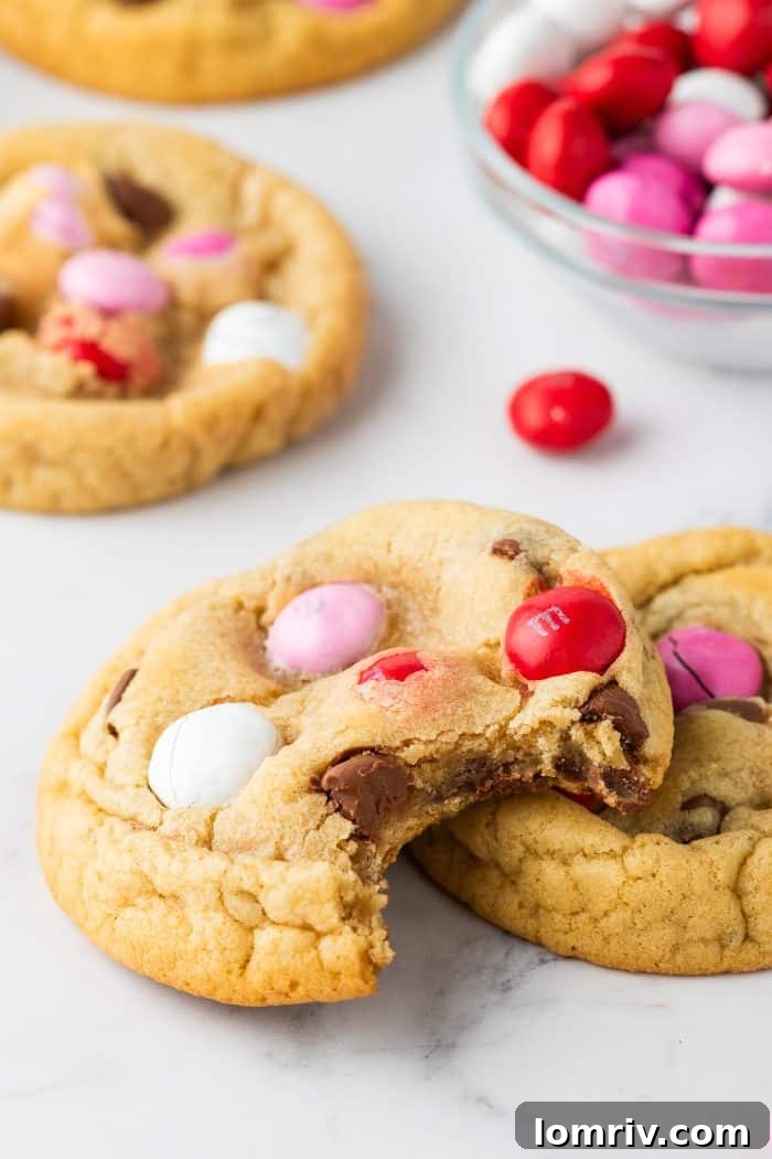 A compelling close-up of a Valentine's M&M cookie with a perfect bite taken out, highlighting its chewy texture and visible candies, with more cookies softly blurred in the background.