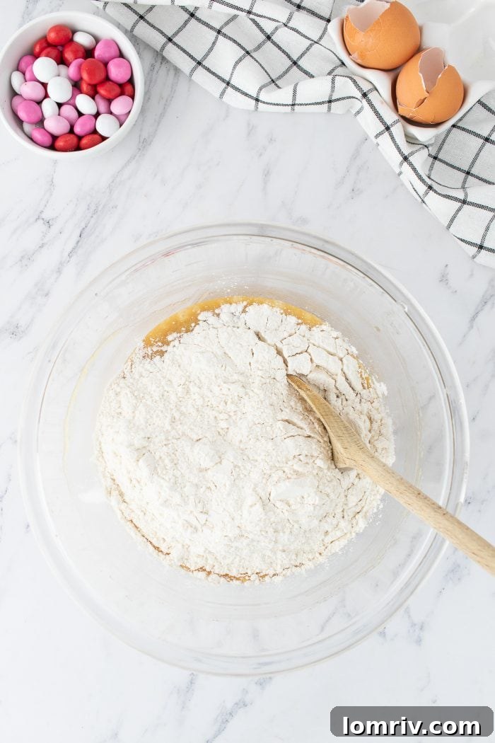 A glass mixing bowl showcases the dough in progress, as the wet and dry ingredients are gently folded together with a wooden spoon to form a cohesive cookie dough.