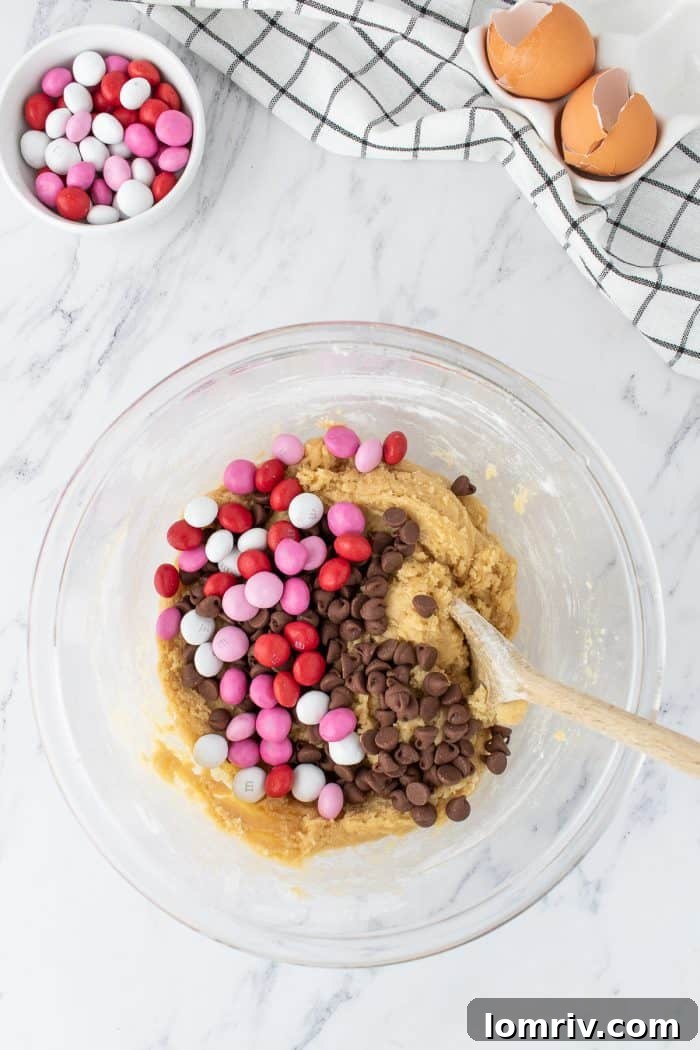 The cookie dough, now richly speckled with colorful M&M's and classic chocolate chips, being gently mixed with a wooden spoon.
