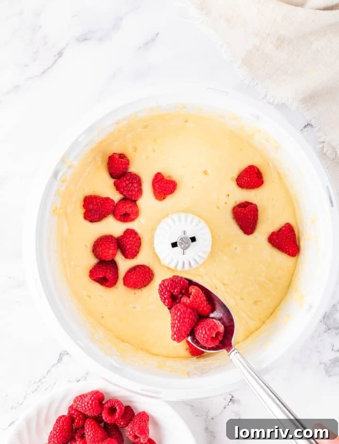 Fresh raspberries being gently folded into the bundt cake batter in a white bowl.
