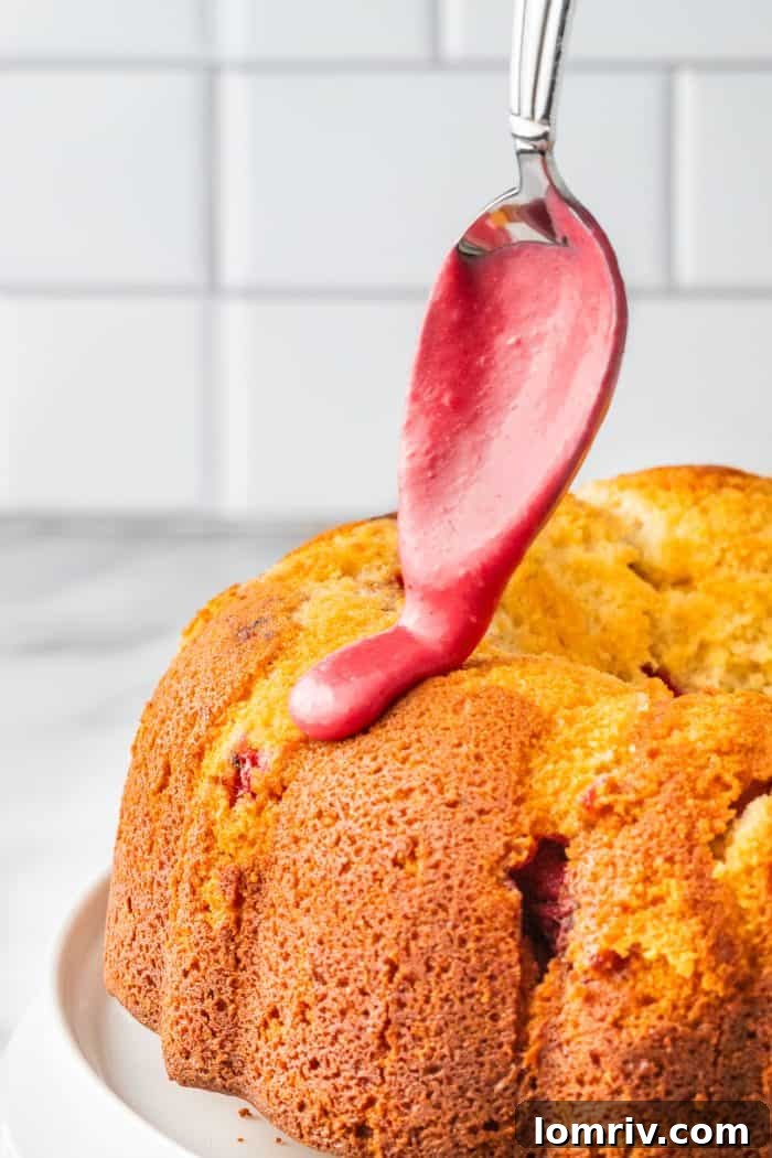 A golden-brown bundt cake being artfully drizzled with the vibrant pink raspberry icing.